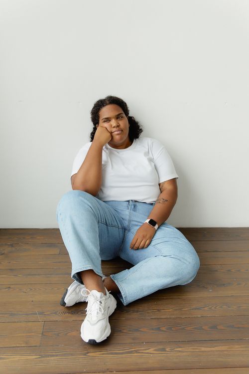Free A confident plus-size woman in denim jeans sitting casually indoors on a wooden floor. Stock Photo