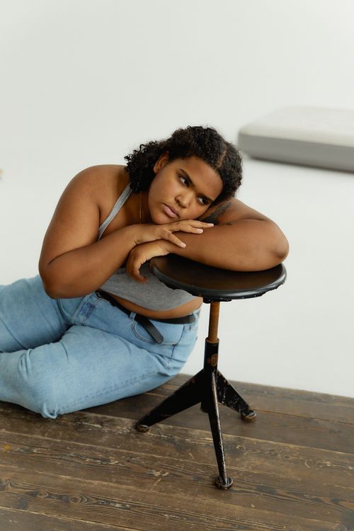 Free Plus-size woman in denim jeans and bra leaning on a stool, expressing body positivity. Stock Photo