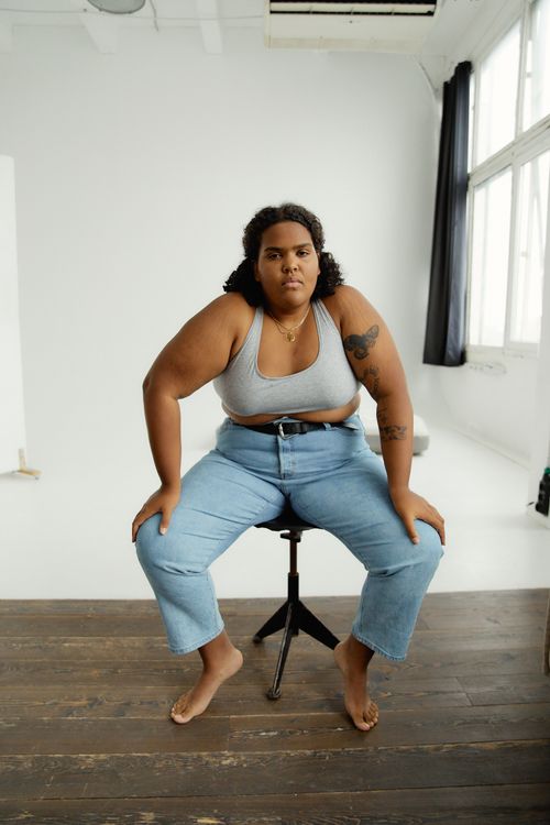 Free Body-positive woman in denim jeans and tank top seated confidently on a stool indoors. Stock Photo