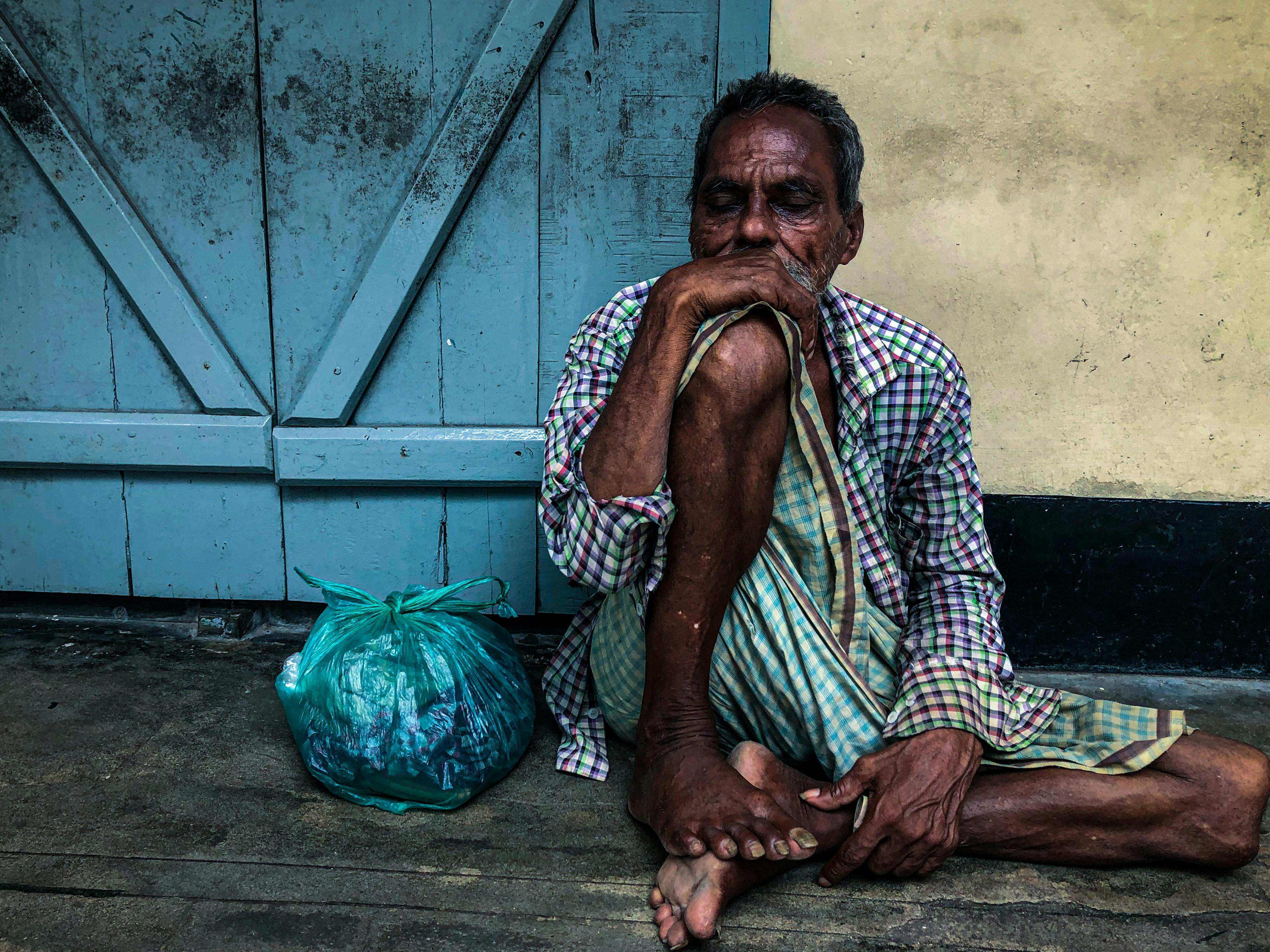 A senior man in plaid shirt and lungi sitting against a blue door with a bag beside him.