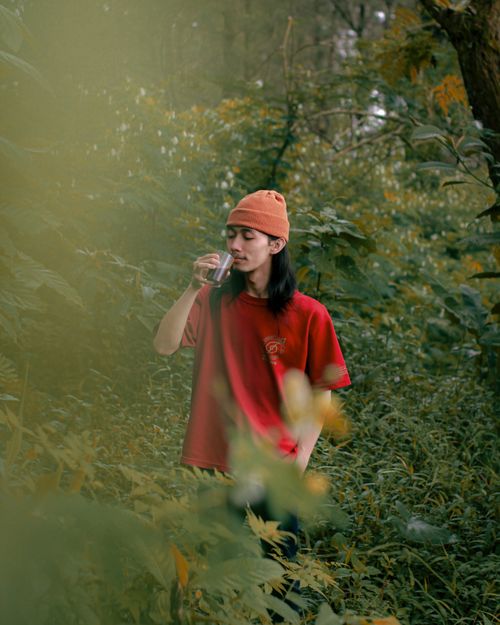 Free A man wearing a red shirt and beanie drinks in a vibrant green forest. Stock Photo