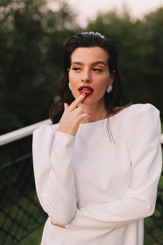Elegant woman in stylish white dress posing outdoors with forest backdrop.