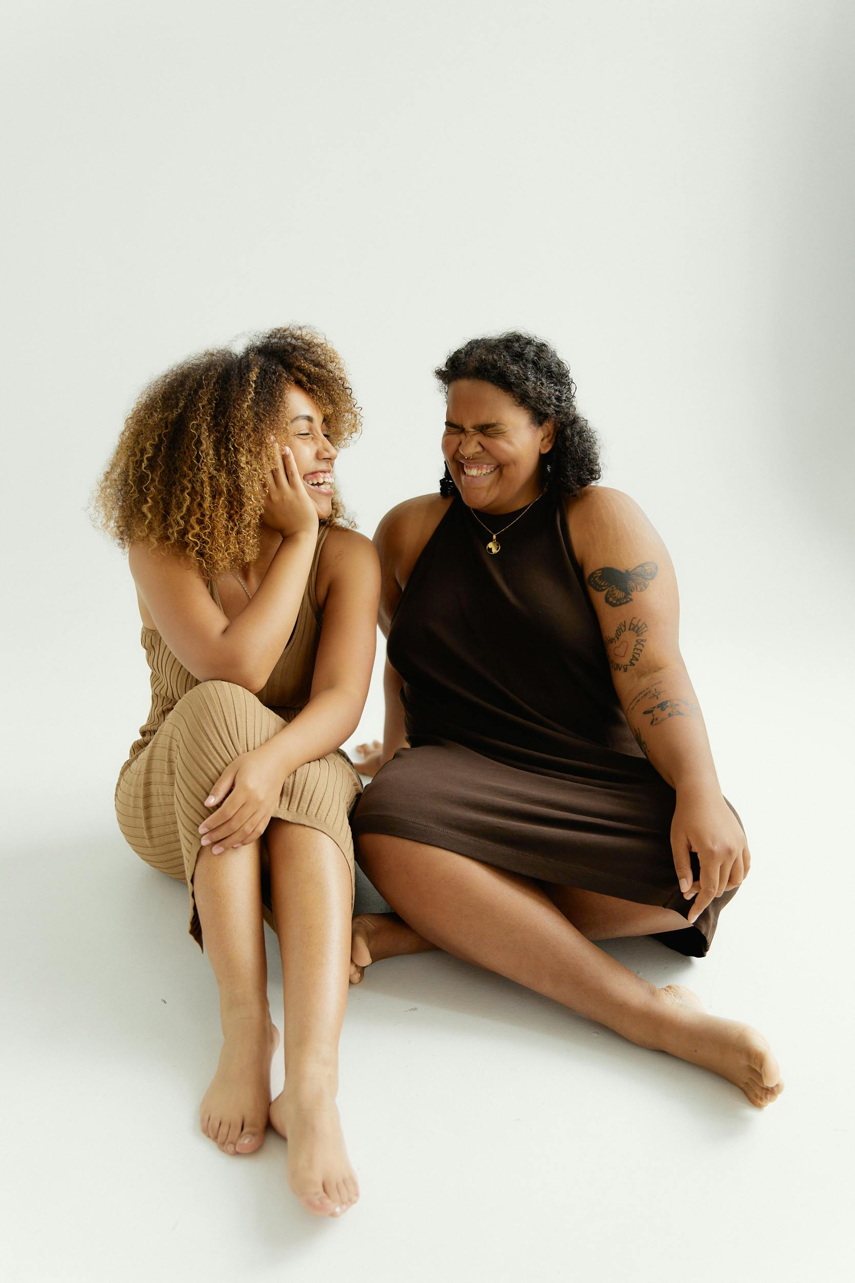 Two women sitting barefoot on a white studio background, smiling and enjoying each other's company.