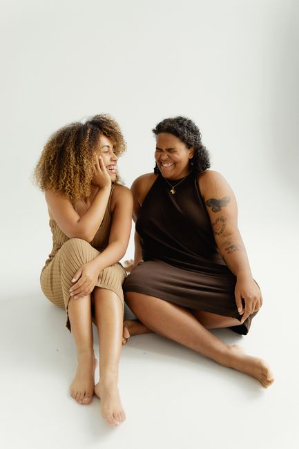 Two women sitting barefoot on a white studio background, smiling and enjoying each other's company.