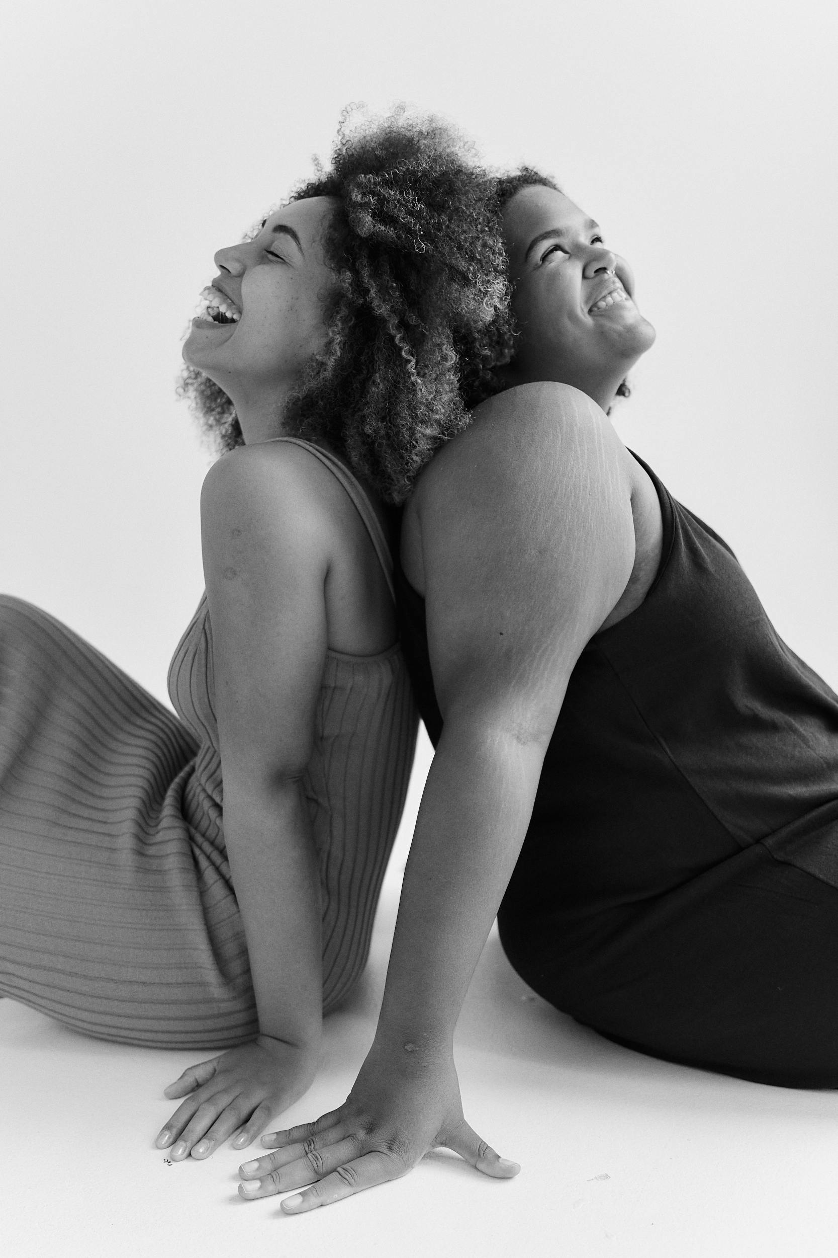 Black and white portrait of two women laughing, sitting back to back in a studio setting.