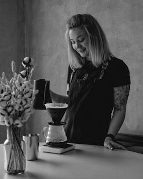 Black and white image of a barista brewing coffee using pour-over technique indoors.