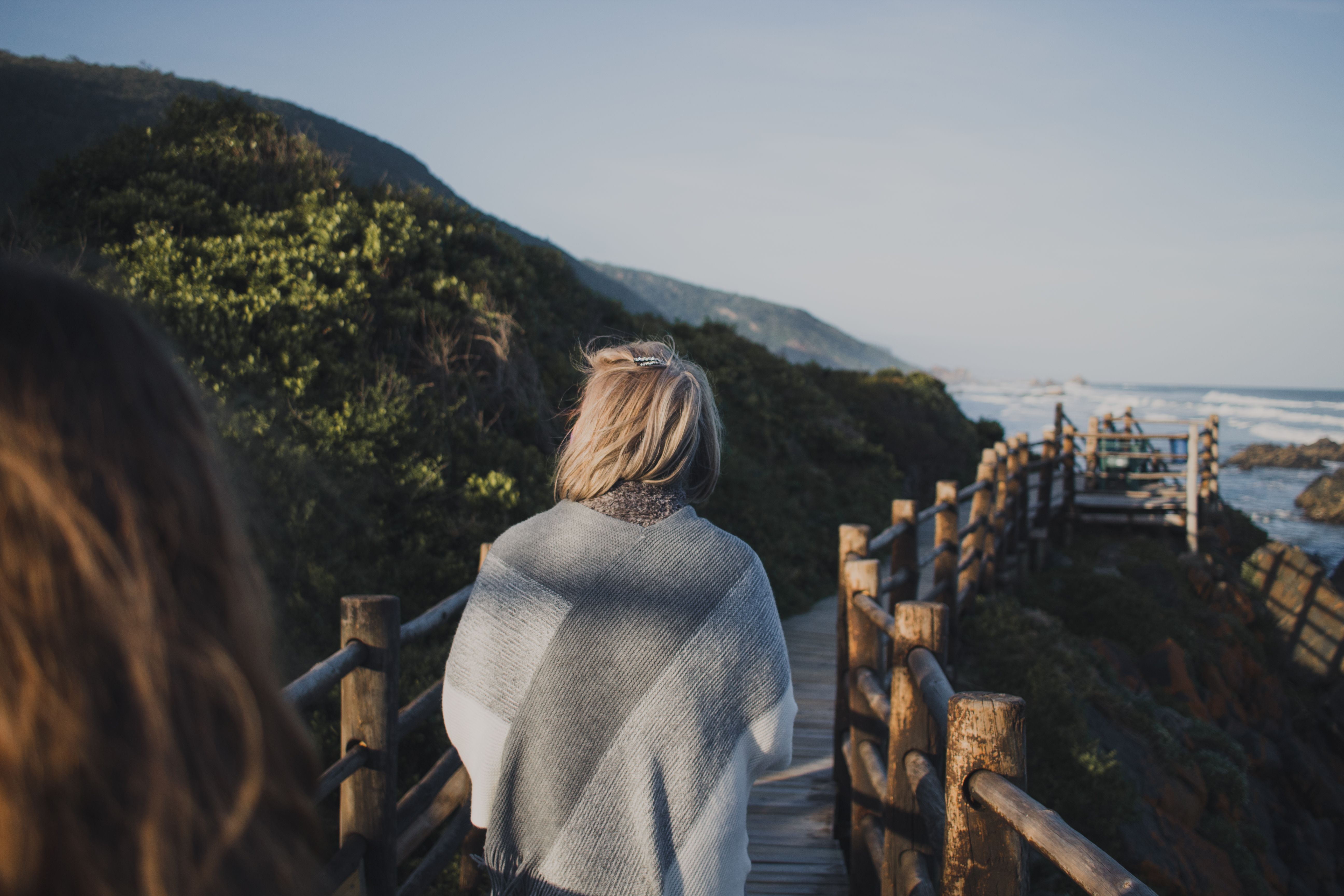 A woman wrapped in a shawl walks along a wooden boardwalk overlooking the ocean.