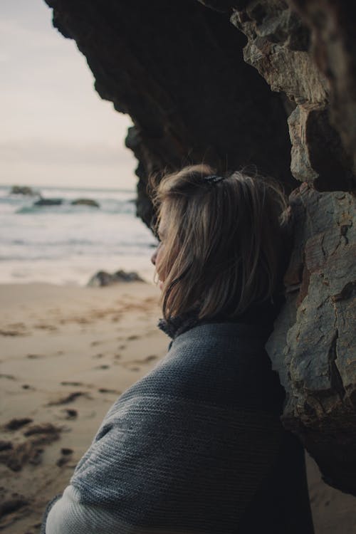 Woman in Gray Sweater Standing beside Gray Rock Formation Near Sea