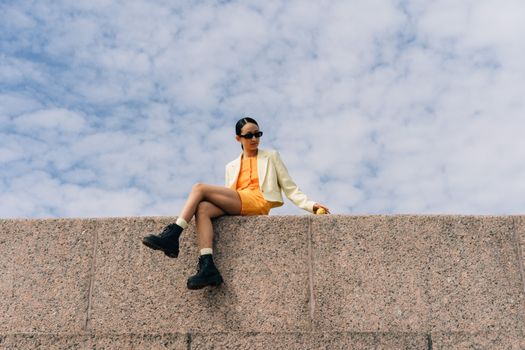 Woman in Sunglasses and Jacket Sitting on Wall