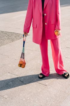 Stylish woman in oversized pink suit holding oranges on a sunlit pavement.