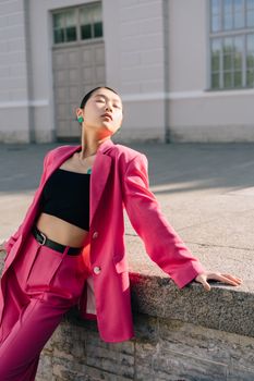 Fashionable young woman in a pink suit poses outside a building, showcasing modern style.