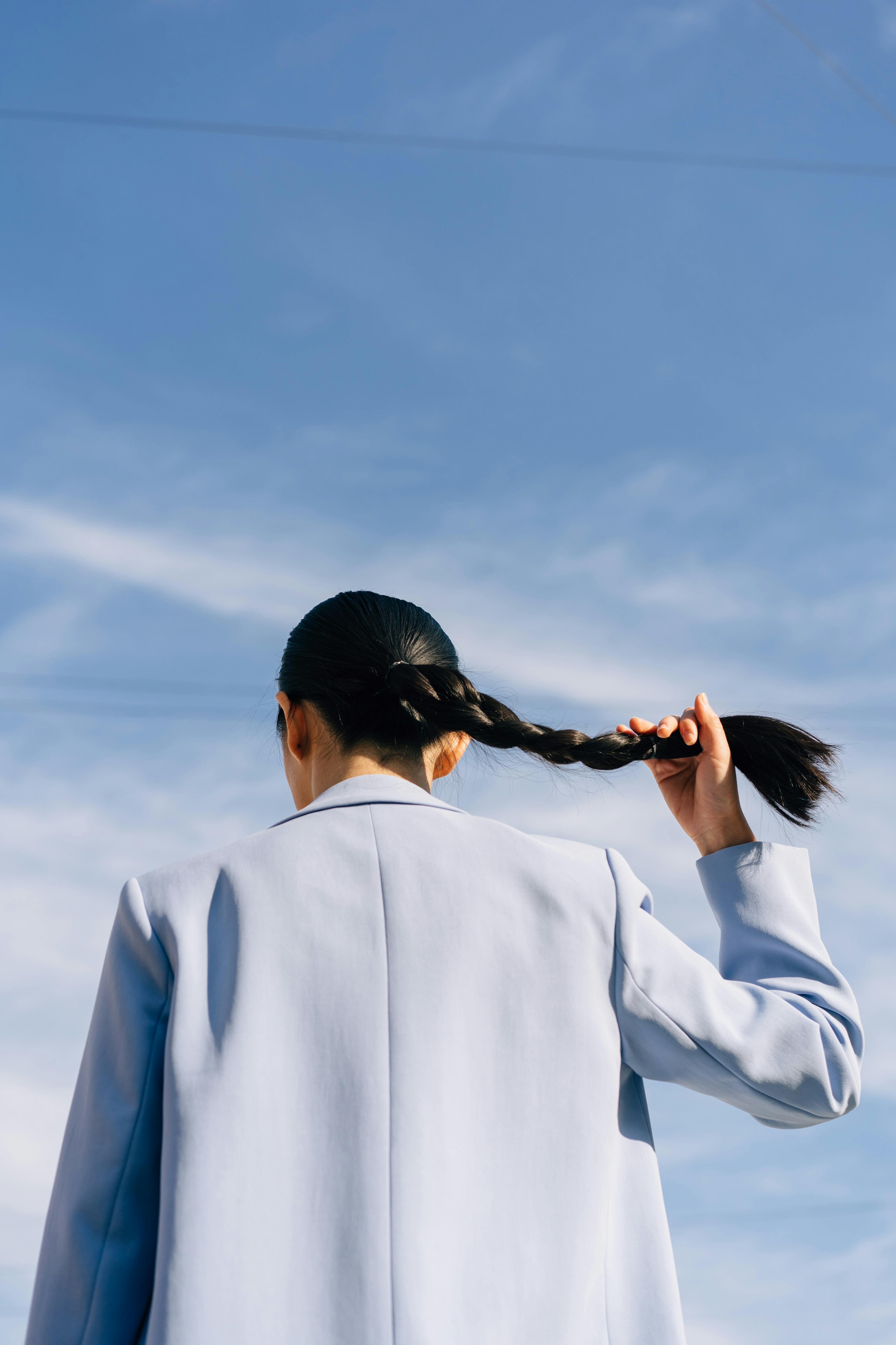 Woman in blue suit holding her ponytail against a clear blue sky, from behind.