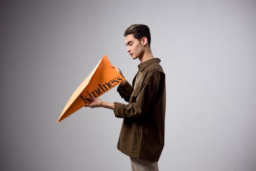 Free A fashion portrait of a male model holding an orange paper airplane with 'kindness' text on a gray background. Stock Photo