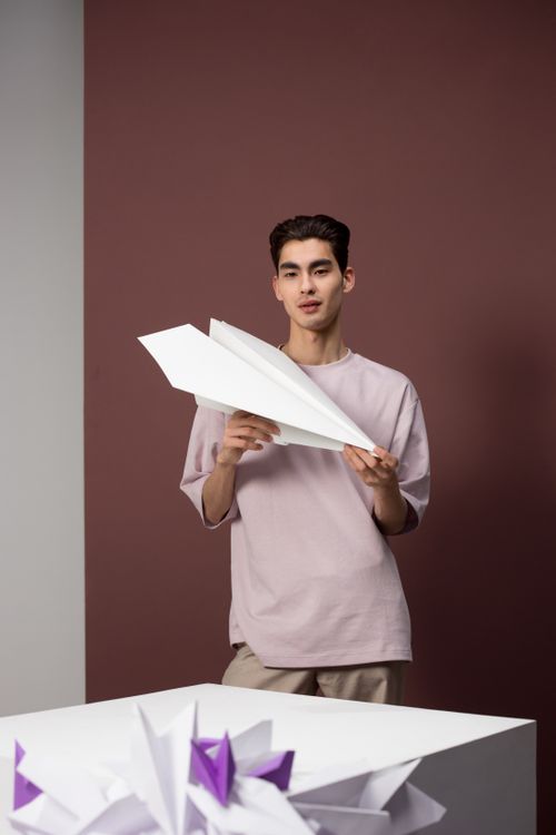 Free A young man stands indoors holding a large paper airplane with creative decor. Stock Photo