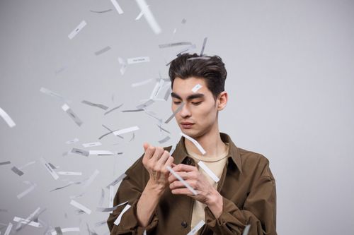 Free Young man standing in a studio amidst falling strips of paper, creating a surreal and creative atmosphere. Stock Photo