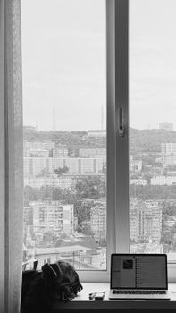 Black and white view of Vladivostok's cityscape, captured indoors through a window with a laptop in the foreground.