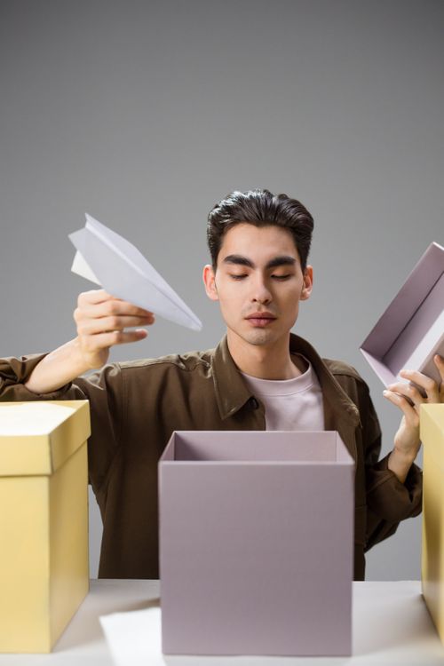Free Portrait of a young adult playing with a paper airplane and boxes indoors, conveying creativity. Stock Photo