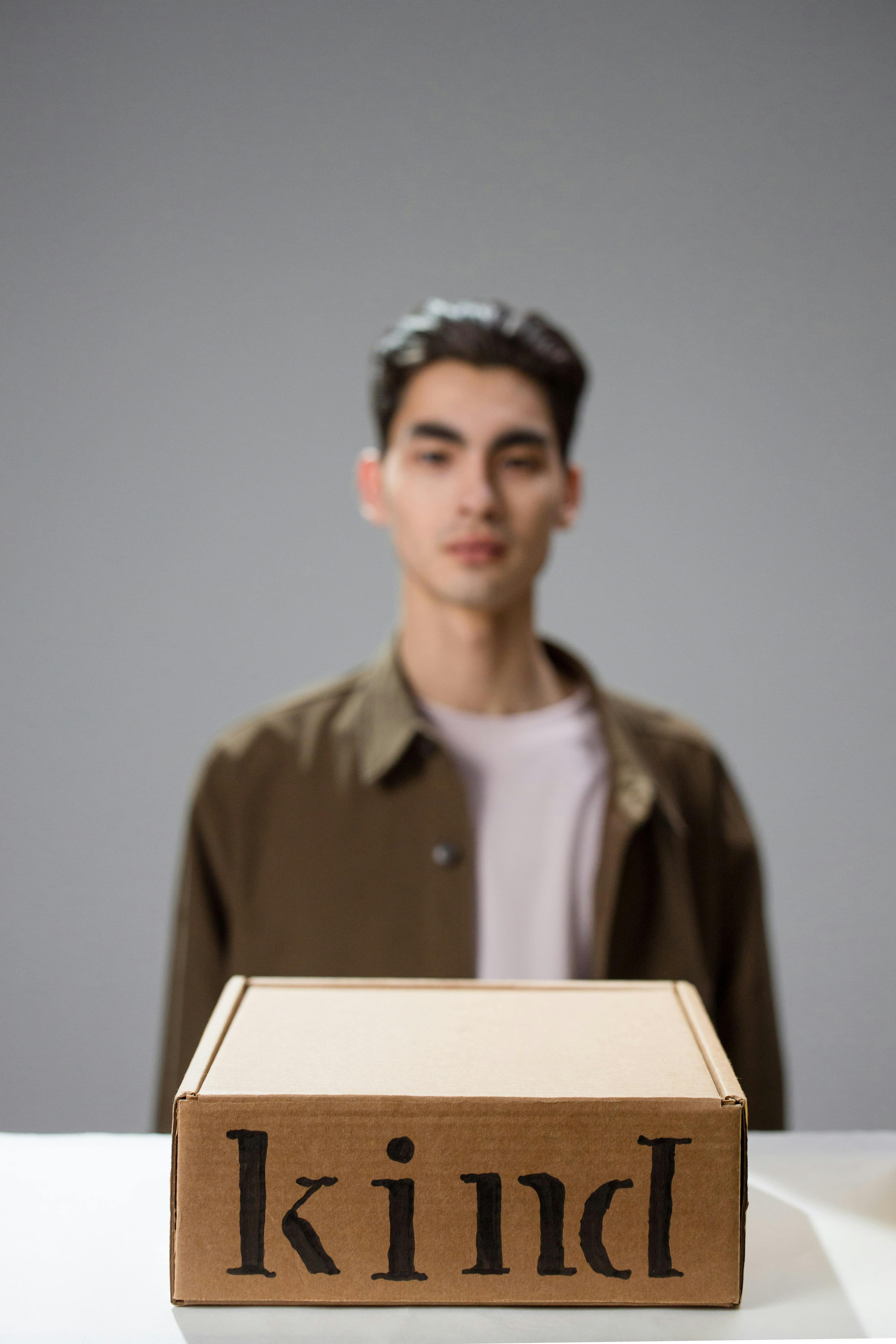 Man in a blurred background with a cardboard box labeled 'kind' on a white surface.