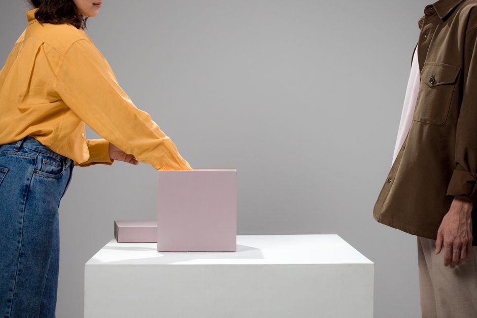 Escena conceptual minimalista de dos personas no reconocibles interactuando sobre una caja en una mesa.