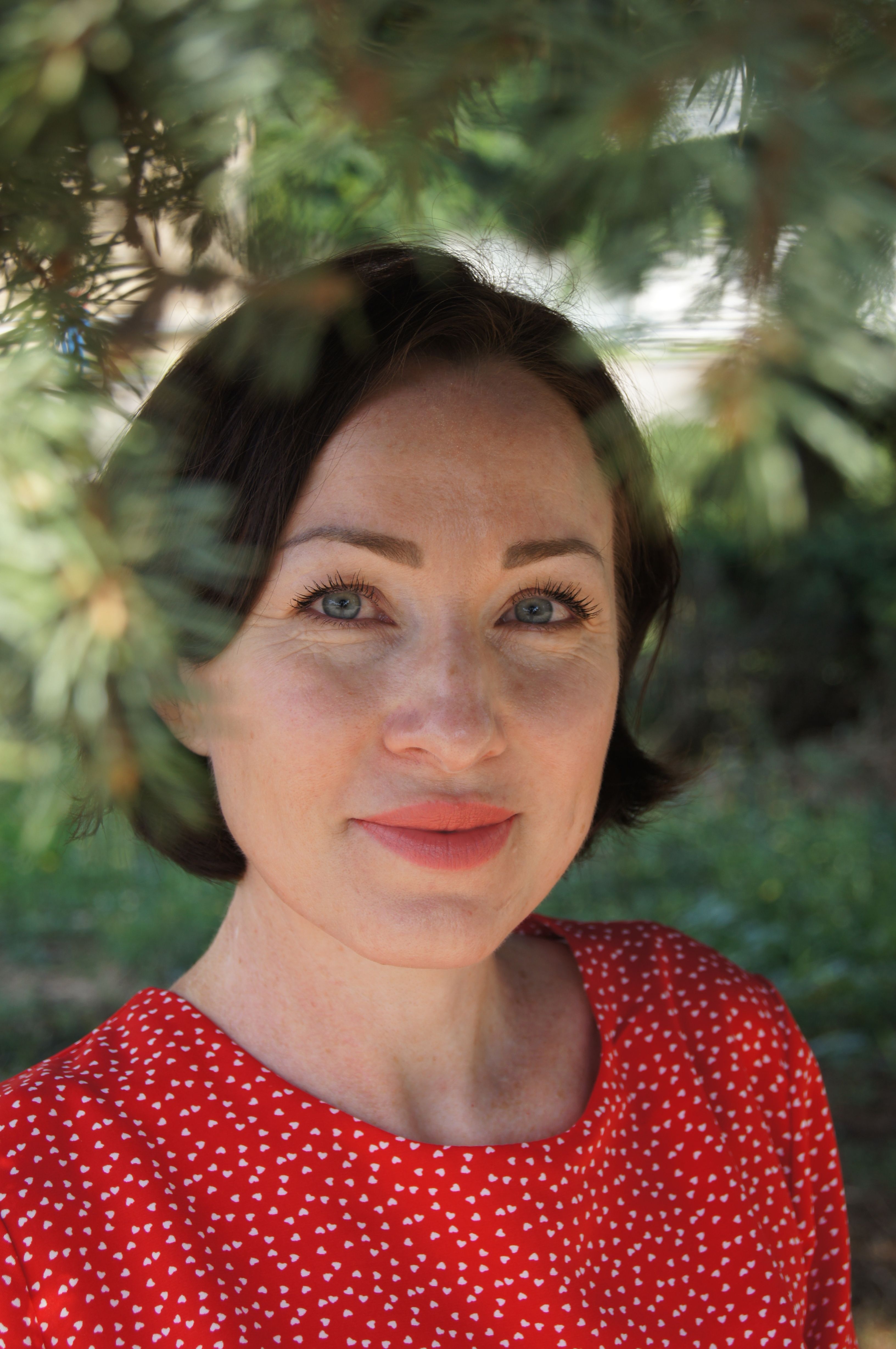 Portrait of a woman with short black hair framed by green leaves, wearing a red dress outdoors.