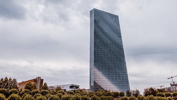 Urban skyscraper towering against a cloudy sky with cityscape and greenery.