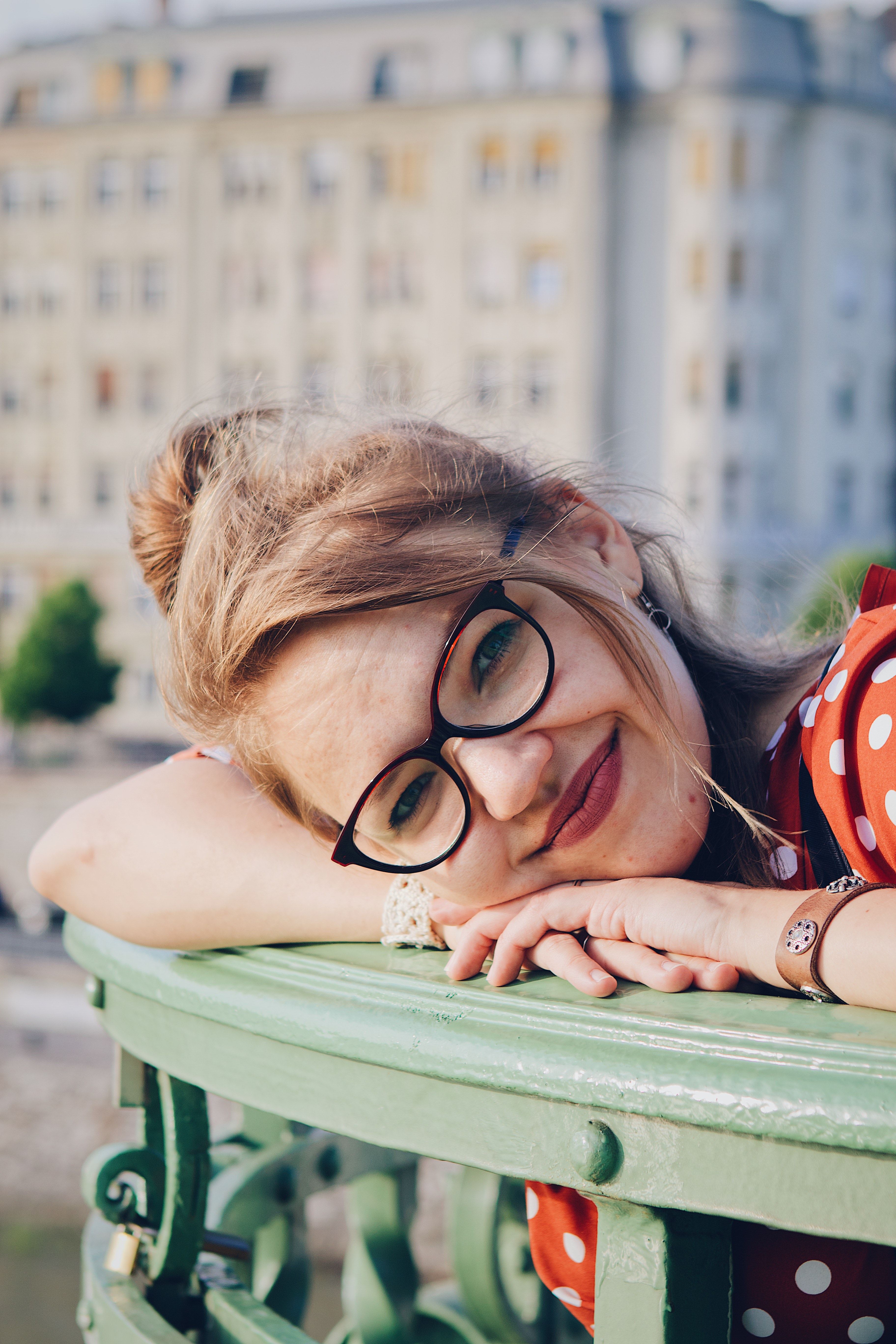 A cheerful woman with glasses leans on a green railing, Budapest in the background.