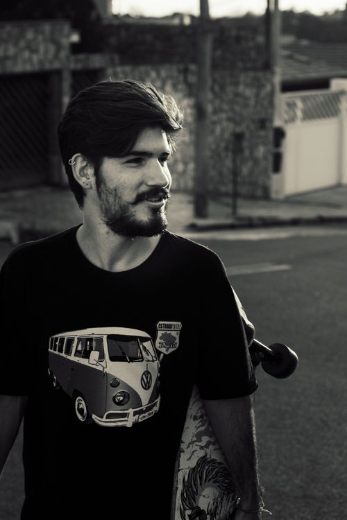 Free A young man with beard holding a skateboard on a city street, wearing a classic van t-shirt. Stock Photo