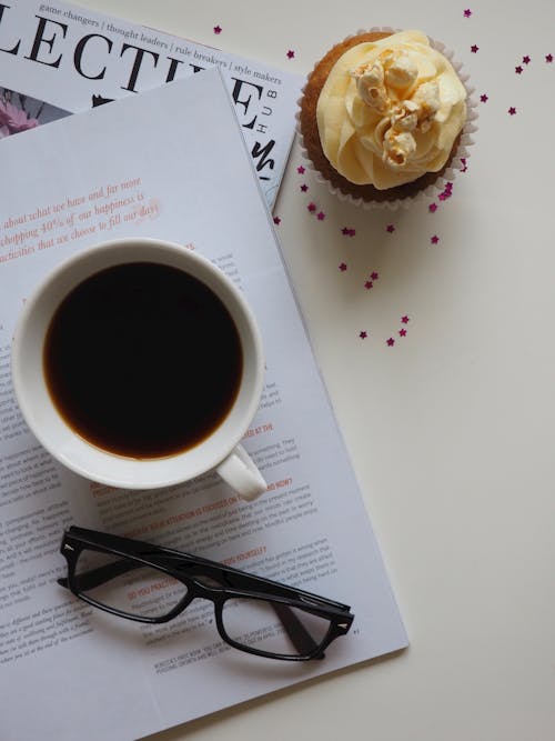 White Ceramic Cup With Coffee on Top of Opened Book and Near Eyeglasses