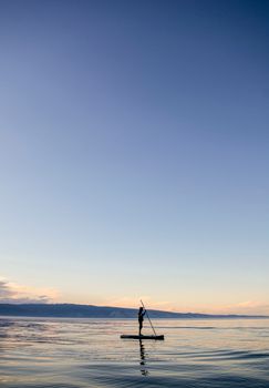 A person paddleboarding on calm ocean waters under a clear sky at sunset.
