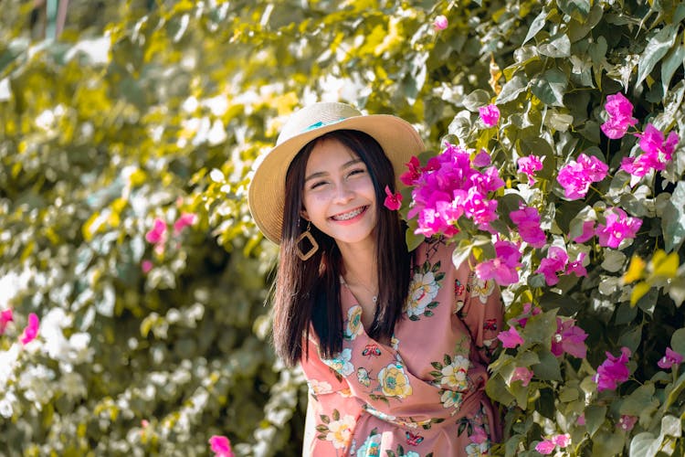 Girl Wearing Floral Dress And Summer Hat Posing With Pink Flower Bush