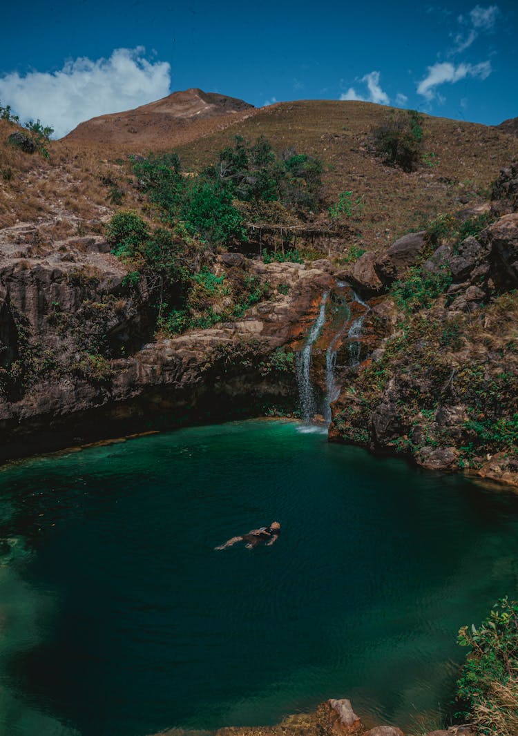 Man Floating In Dark Turquoise Water And Beige Mountain Landscape