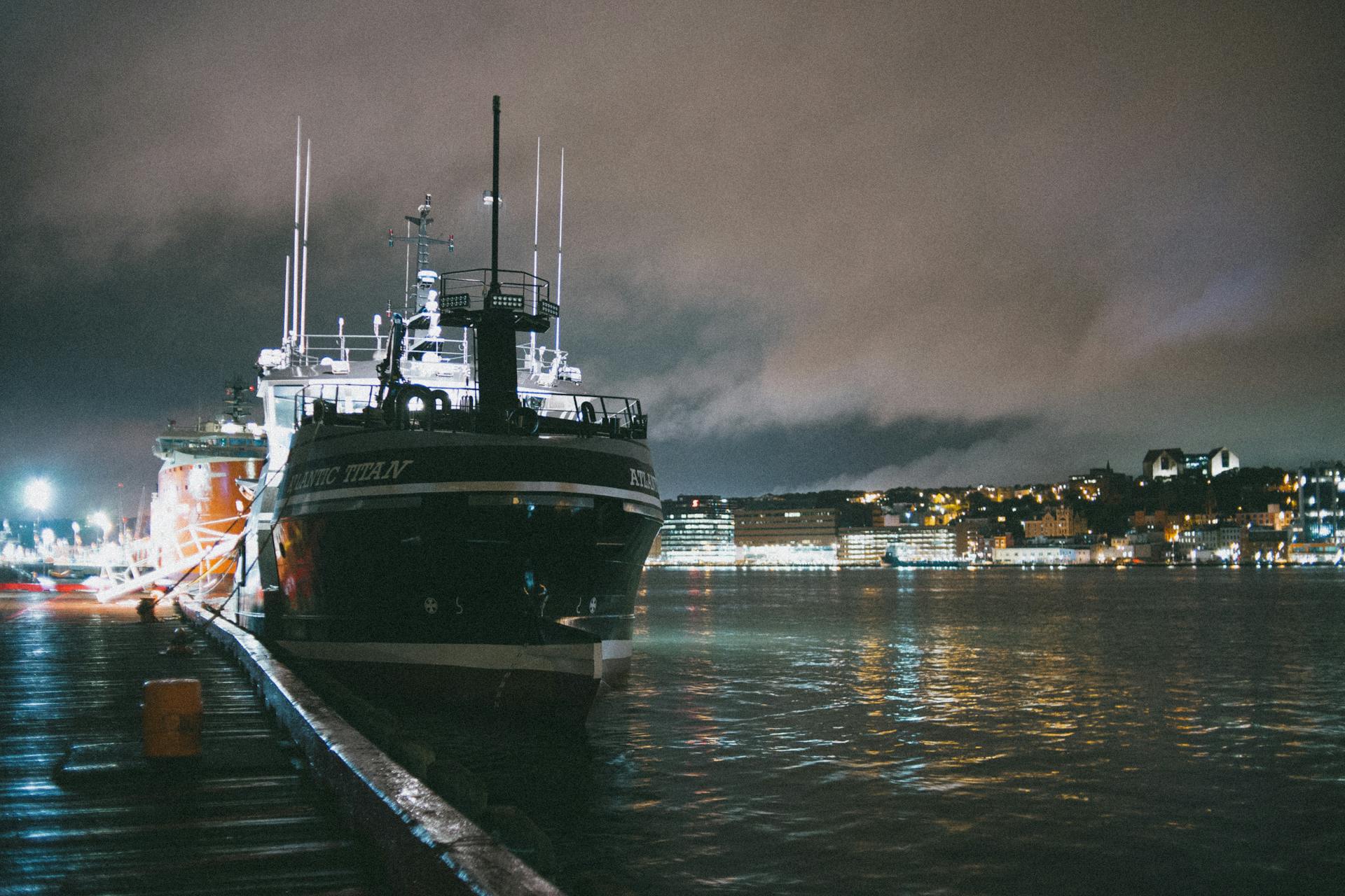 A large ship docked at a city harbor during nighttime, with city lights reflecting on water.