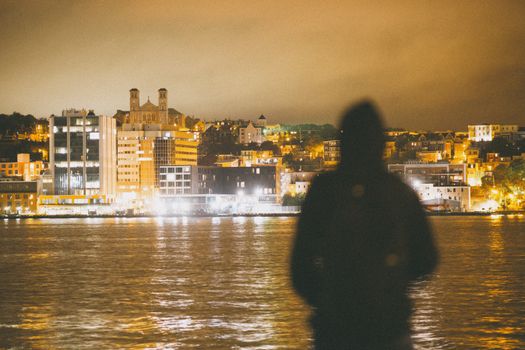 A silhouette of a person standing by a river, overlooking an illuminated city waterfront at night.