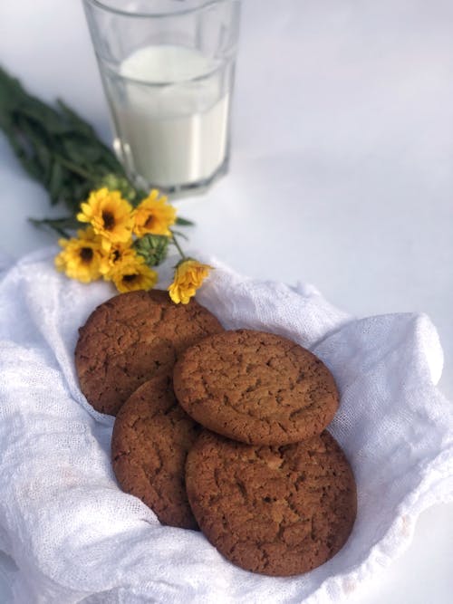 Brown Cookies on White Table Napkin Beside a Glass of Milk and Yellow Flowers