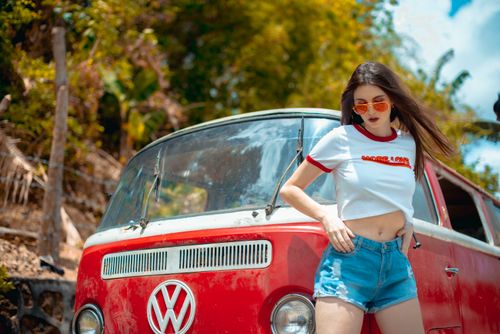 Free Young woman in denim shorts posing by a classic red Volkswagen van outdoors. Stock Photo