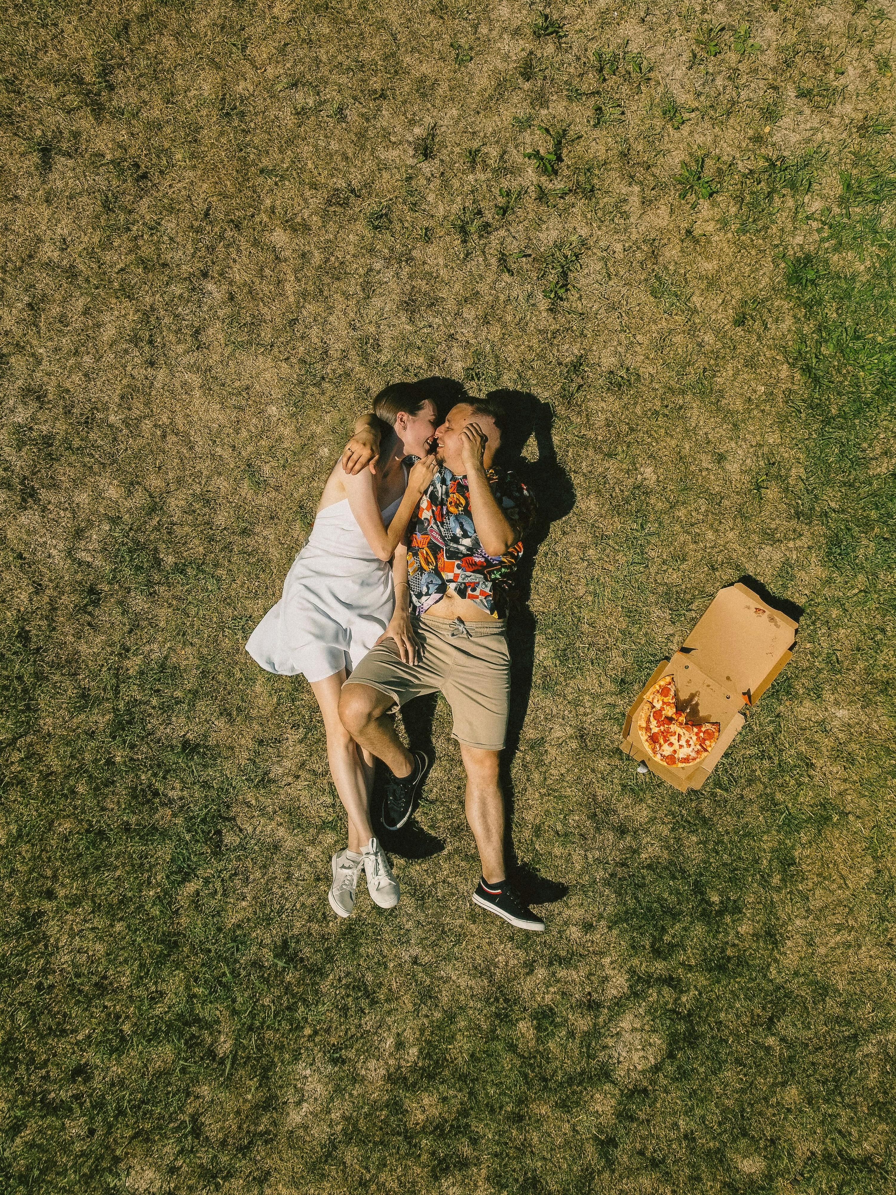 Couple lying on grass with pizza box, enjoying a sunny day outdoors.