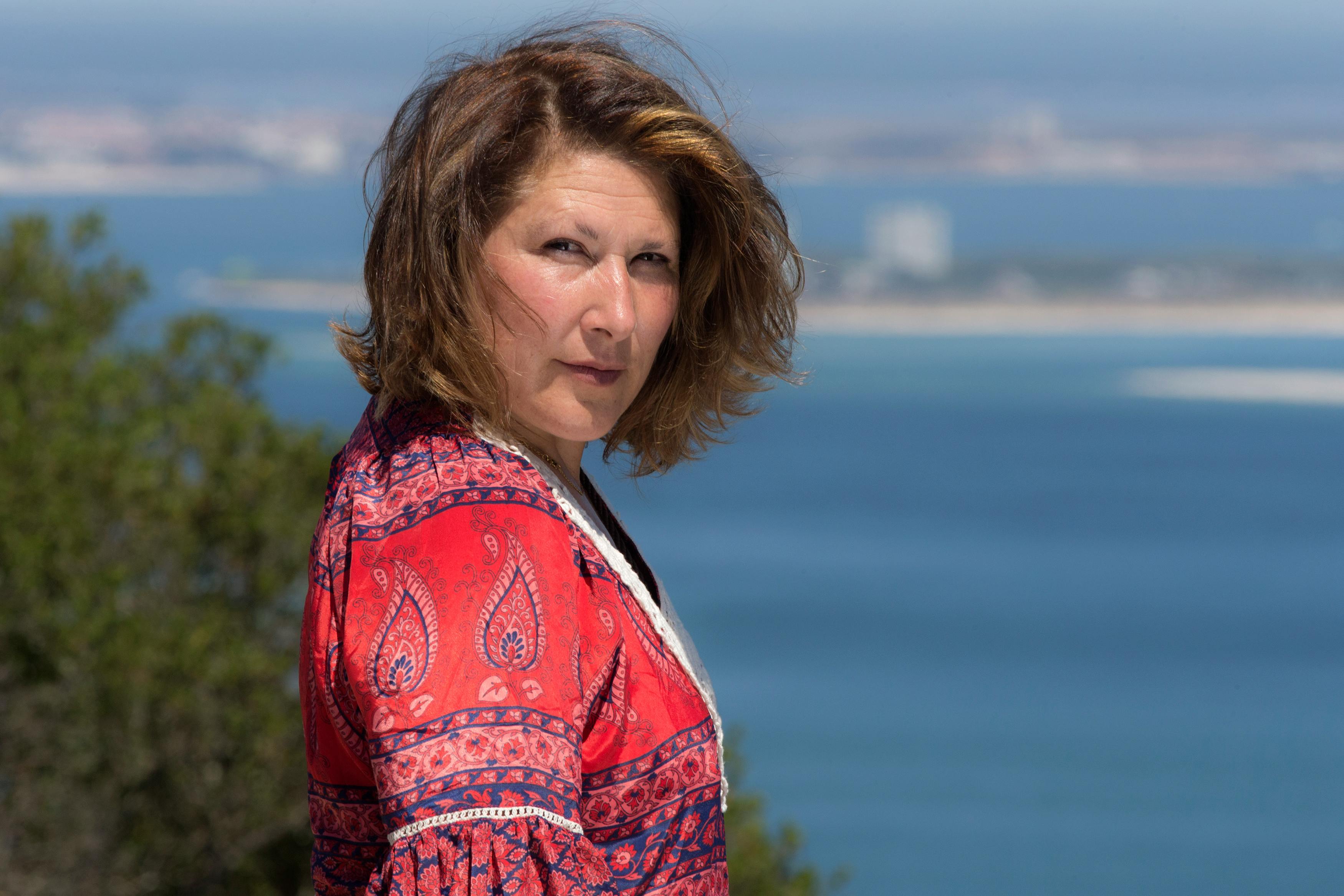 Woman with short hair in a red top standing outdoors with ocean view.
