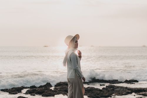 Free A woman in a hijab and hat strolls along a rocky seashore during sunset, capturing a moment of peace. Stock Photo
