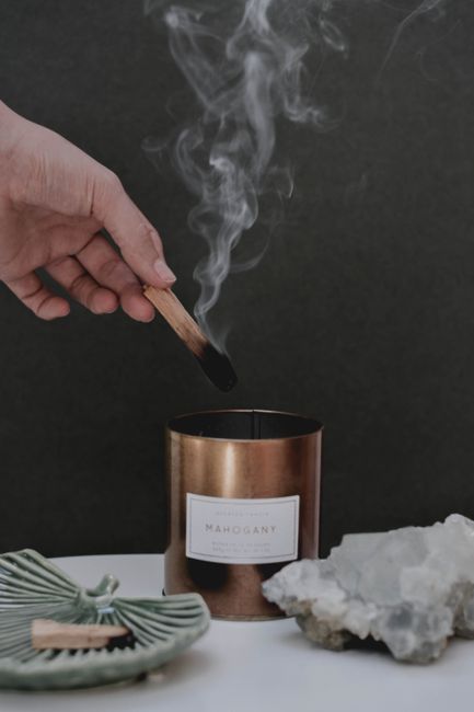 A hand lighting a mahogany candle with a burning palo santo stick, surrounded by crystals.