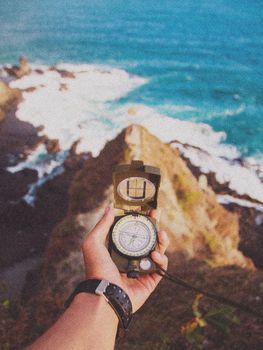 A hand holds a compass overlooking a rocky coastline and ocean in Jogja, Indonesia.
