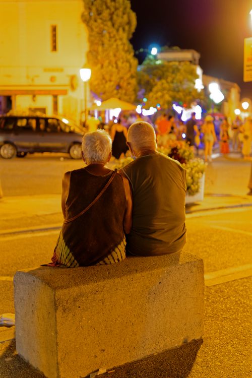 Free An elderly couple sitting on a concrete bench in a vibrant nighttime street scene in Marseille. Stock Photo