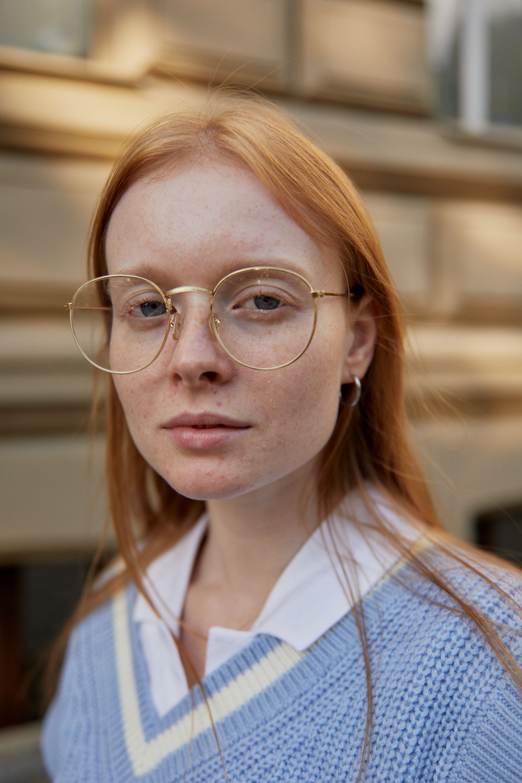 Close-up portrait of a young woman with eyeglasses, wearing a blue sweater outdoors.