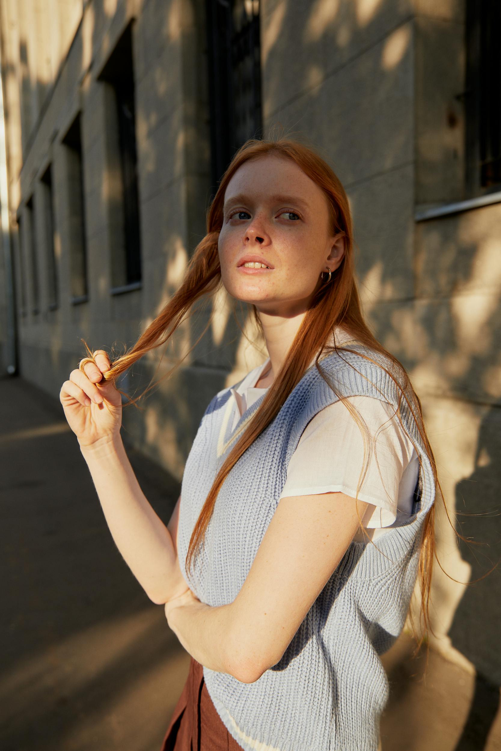 A young woman with red hair poses thoughtfully on a sunlit urban street, wearing a sweater vest.