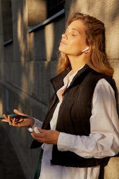 A woman in a black vest enjoys music with earphones in the warm sunshine outdoors.
