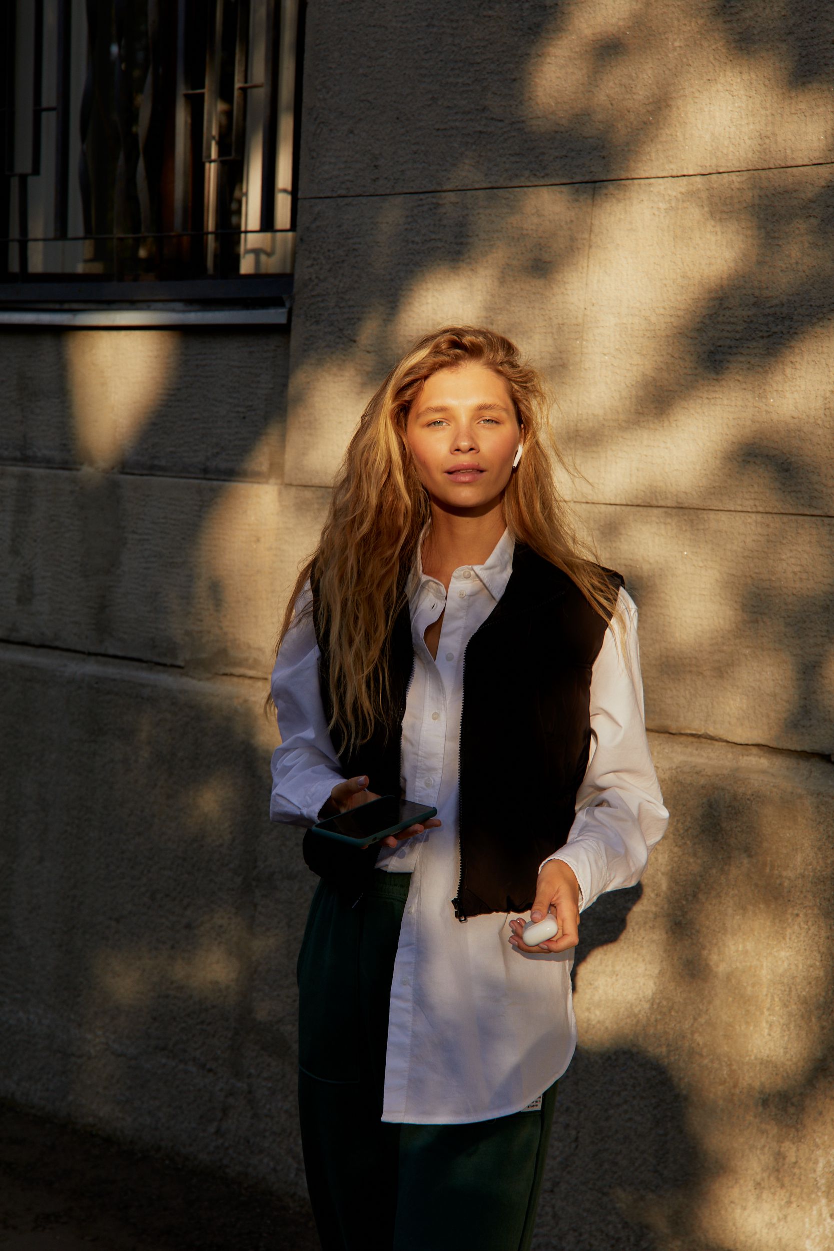 Woman holding phone and earphones, standing by a concrete wall in sunlight.