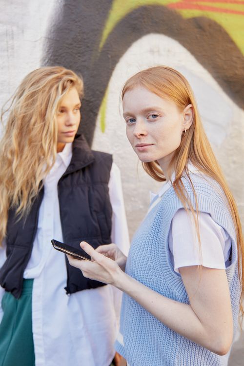 Free Two stylish young women standing outdoors, one holding a smartphone, against a vibrant graffiti background. Stock Photo