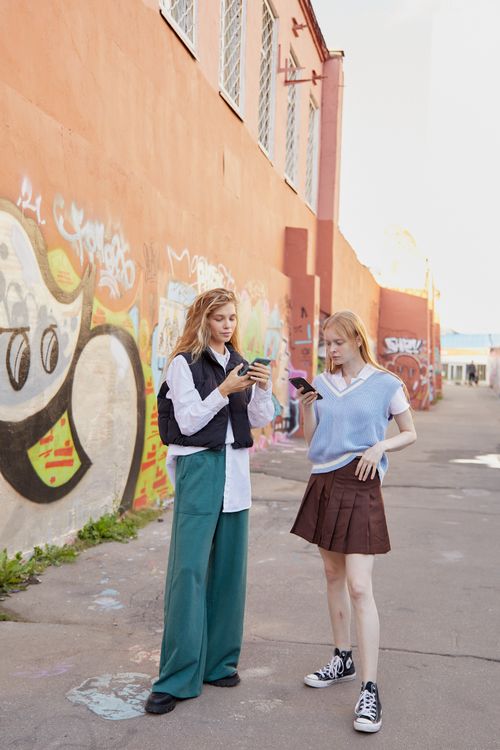 Free Two fashionable teenagers using smartphones in an urban graffiti setting, showcasing modern youth culture. Stock Photo