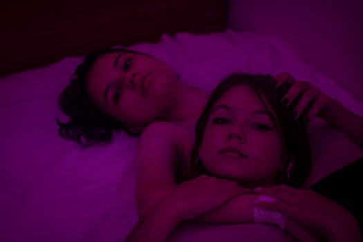 Two women lying closely on a bed illuminated by soft pink lighting, creating a warm and intimate atmosphere.