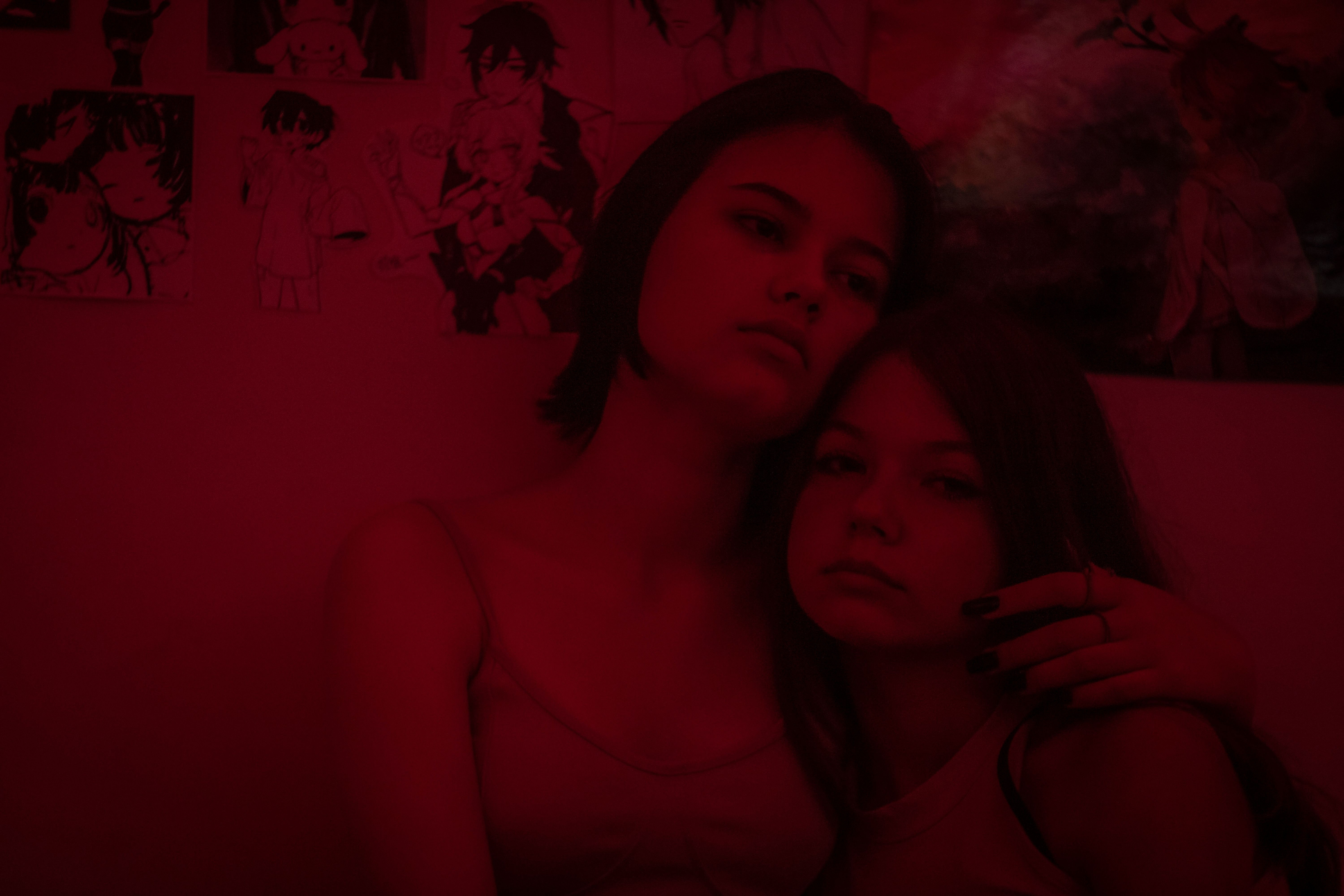 Two women share a tender moment under a red glow surrounded by anime artwork.