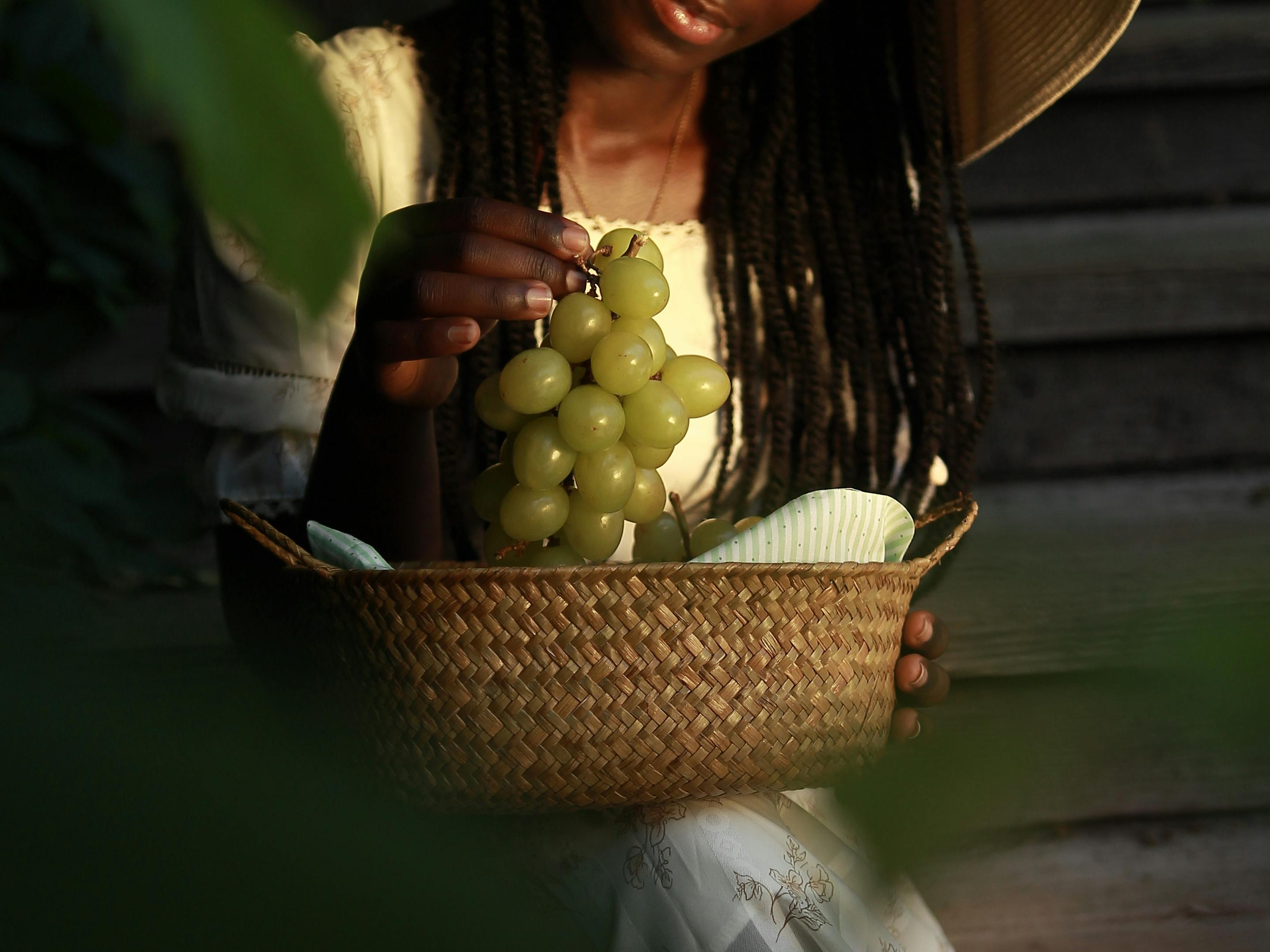 A woman enjoying a sunny day, holding a cluster of green grapes in a woven basket.
